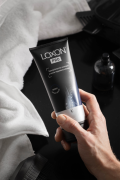 Czarna tuba LOXON PRO szamponu przeciw wypadaniu włosów, trzymana w dłoni. W tle biały ręcznik i grzebień.