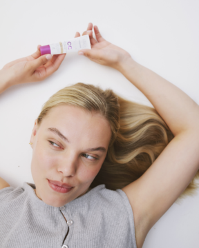 Kobieta z blond włosami leży, trzymając nad głową tubkę korygującego kremu CC Lumene SPF 20, wyrównującego koloryt.