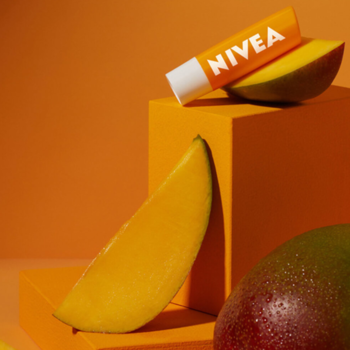 NIVEA Mango Shine pomadka w sztyfcie, na tle plasterków mango i pomarańczowych bloków. Długotrwałe nawilżenie ust.