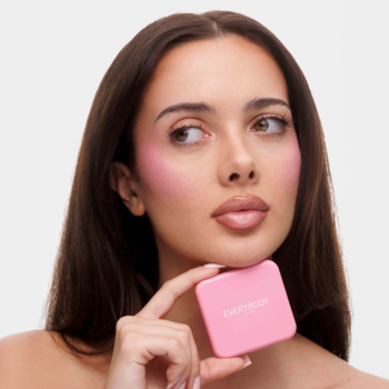 Róż satynowy Everybody London Lily's Doll. Kobieta z różowymi policzkami trzyma różową kwadratową puderniczkę.