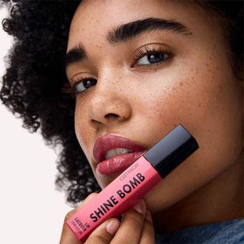 Błyszczyk Catrice Shine Bomb Flirt Alert w różowej tubce, trzymany przez kobietę z ustami w koralowym różu.