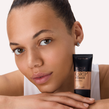 Kobieta prezentuje rozświetlającą bazę Catrice Bronze Glow Grip Primer Sun Glazed, brązową tubkę przy twarzy.