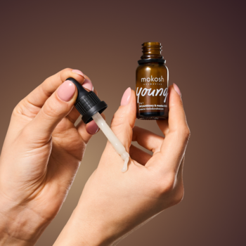 Mokosh Young serum przeciwstarzeniowe w brązowej butelce z pipetą, aplikowane na dłoń. Zbliżenie na kroplę produktu.