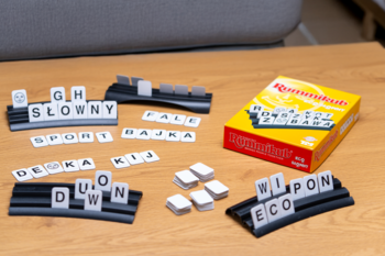 Zestaw gry Rummikub Eco Słowny rozłożony na drewnianym blacie, z tekturowymi literami i czarnymi podstawkami.