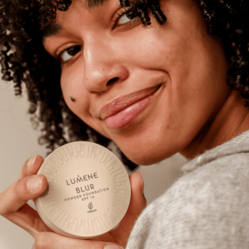 Uśmiechnięta kobieta z kręconymi włosami, prezentuje jasnobeżowy puder Lumene Blur SPF 15.