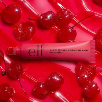 Różowy balsam do ust e.l.f. Glow Reviver Melting Wild Cherry leży wśród czerwonych wiśni na lśniącym, czerwonym tle.