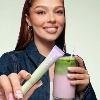 Uśmiechnięta kobieta wyciąga balsam Everybody Peptide Lip Balm Lavender Matcha w zielono-fioletowej tubce, obok napój.