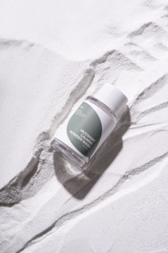 Isntree Mugwort Calming Powder Wash, transparentna butelka z zieloną etykietą, leży na falistym białym proszku.