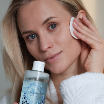 Kobieta aplikuje kojącą wodę micelarną Lumene Herkkä na twarz wacikiem. Przejrzysta butelka z niebieską etykietą w dłoni.