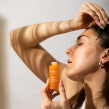 Kobieta aplikuje rozświetlającą esencję Lumene Nordic-C Valo z pomarańczowej butelki na policzek.