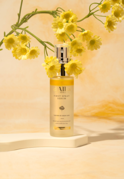 d'Alba White Truffle First Spray Serum w przezroczystej butelce z żółtą cieczą, na białej platformie z żółtymi kwiatami.