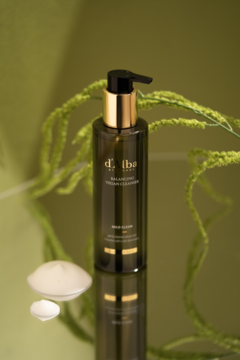 Wegański żel d'Alba Balancing Cleanser, ciemna butelka z pompką na lustrze. Obok biała pianka, zielone liście w tle.
