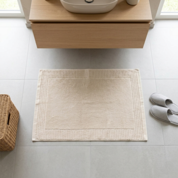 Kremowy, prostokątny dywanik łazienkowy z delikatnymi pasami na jasnych płytkach. Widok z góry.