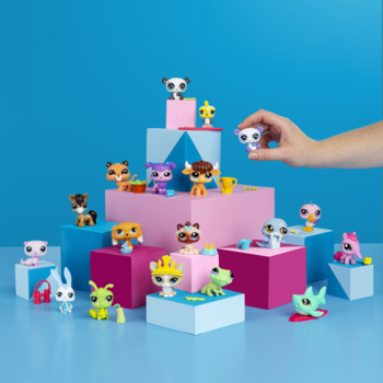 Kolorowe figurki Littlest Pet Shop Generacji 7 na różowo-niebieskich klockach; ręka trzyma jedną figurkę z dużymi główkami.