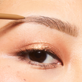 Zbliżenie na oko, brwi korygowane jasnobrązową kredką, tworzącą naturalne włoski. Na powiece złoty cień.