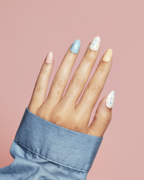 Ręka z naklejkami na paznokcie Ardell Nail Addict, pastelowe: błękitne, żółte, różowe i w kropki, na różowym tle.