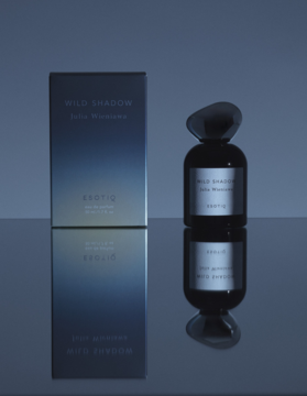 Perfumy Wild Shadow Exotic: ciemna butelka z unikalnym korkiem i pudełko z gradientem, odbite w tafli.