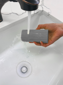 Szary, prążkowany czyścik lub gąbka kuchenna, płukany pod bieżącą wodą w białym zlewie. Widok z góry.