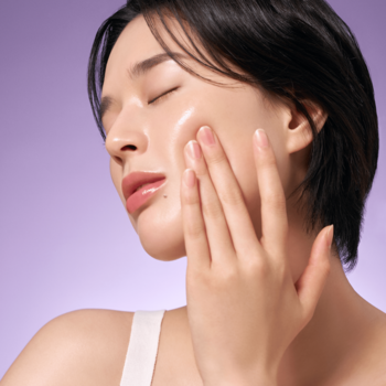 Zbliżenie na kobietę z gładką cerą, delikatnie wmasowującą w skórę nawilżające serum z bakuchiolem na lawendowym tle.