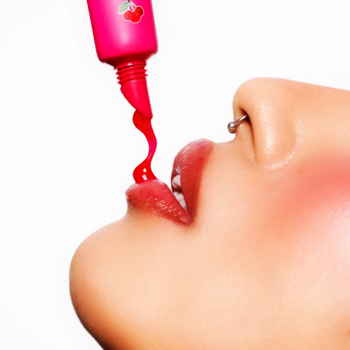 Zbliżenie: różowy Fluff Candy Lip Balm Cherry Crush wyciskany z tubki prosto na otwarte usta modelki.