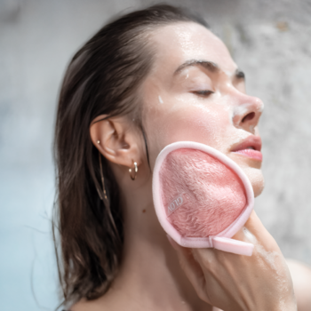 Kobieta z zamkniętymi oczami myje twarz różowym, puszystym padem do demakijażu, widok z profilu.