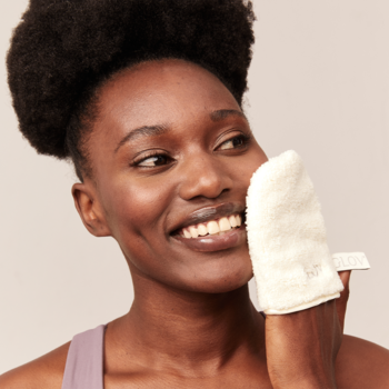 Uśmiechnięta kobieta z ciemnymi włosami delikatnie oczyszcza twarz puszystą rękawicą do demakijażu.