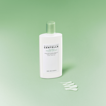 Biała butelka SKIN1004 Madagascar Centella Tea-Trica Soothing Sun Milk SPF 50+ dla cery trądzikowej, smugi kremu obok, tło jasnozielone.