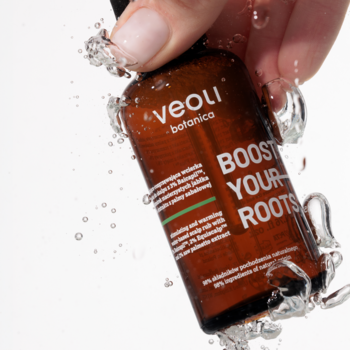 Ręka trzyma brązową butelkę wcierki wodnej Veoli Botanica Boost Your Roots, wokół rozpryskująca się woda.