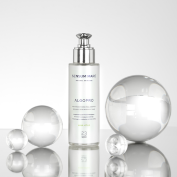 Biała butelka esencji mikrozłuszczającej Algopure z pompką, otoczona szklanymi kulami, na białym tle.