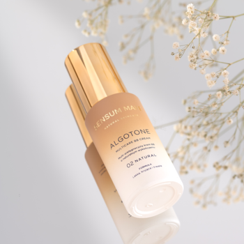 Krem BB Sensum Mare Algotone 02 Natural, złota butelka z gradientem, odbita na lustrze, kwiaty w tle.