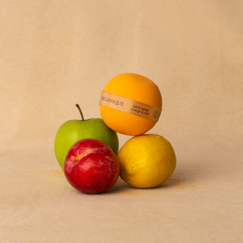 Pomarańczowa kula kąpielowa z etykietą, obok zielonego jabłka, czerwonej śliwki i żółtej cytryny na beżowym tle.