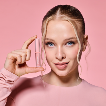 Kobieta w różowej bluzce trzyma matową pomadkę Everybody London Velvet Matte Dusty Silk. Różowe tło, zbliżenie na produkt.