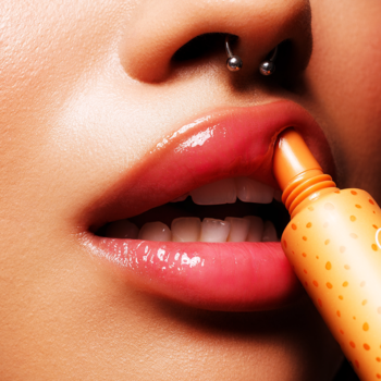 Zbliżenie na lśniące usta z piercingiem, aplikujące Fluff Candy Lip Balm o zapachu ciasteczek z pomarańczowej tubki.