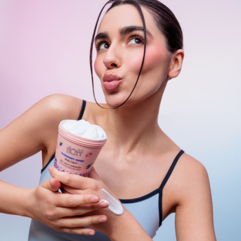 Kobieta nakłada Fluff Blueberry Mochi Body Yogurt na ramię. Różowy słoiczek z białym kremem na pastelowym tle.