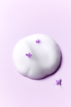 Biała, puszysta pianka oczyszczająca z fioletowymi płatkami na jasnoliliowym tle. Widok z góry.