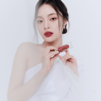 Matowy tint Muzigae Mansion Spicy, trzymany przez kobietę z czerwonymi ustami na jasnym, rozmytym tle studyjnym.