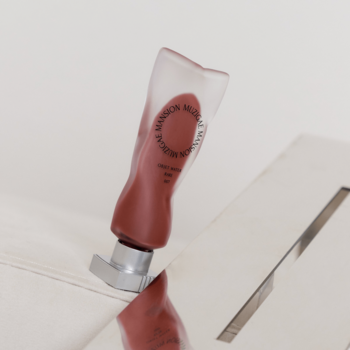 Muzigae Mansion Rare matowy tint do ust, transparentna tubka w odcieniu brązowo-czerwonym, na jasnej powierzchni.