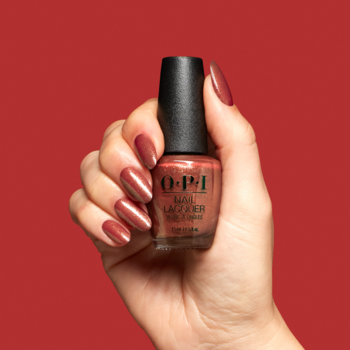 Dłoń trzyma butelkę lakieru OPI Ginger Snapped, ciepły brąz z miedzianym wykończeniem. Pomalowane paznokcie na czerwonym tle.