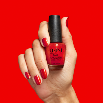 Dłoń z jaskrawoczerwonymi paznokciami trzyma butelkę błyszczącego lakieru OPI Spread the Cherry. Widok na czerwonym tle.