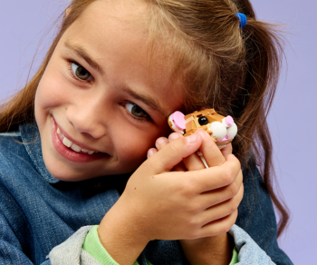 Uśmiechnięta dziewczynka z figurką chomika LEGO Creator 3 w 1 blisko twarzy, na fioletowym tle.