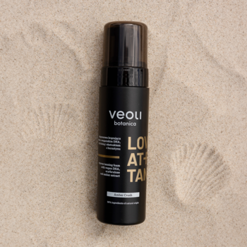 Czarna butelka ekspresowej pianki brązującej Veoli Botanica Love At First Tan Amber Crush na piasku, widok z góry.