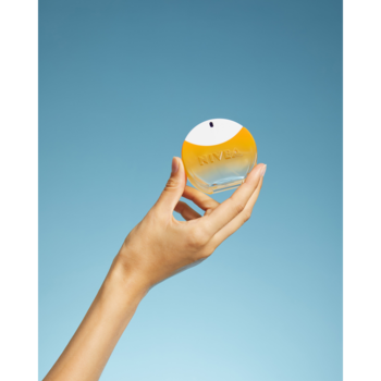 Damska dłoń trzyma okrągłą butelkę perfum SUN GOLD z żółtym płynem i białym korkiem na jednolitym błękitnym tle.