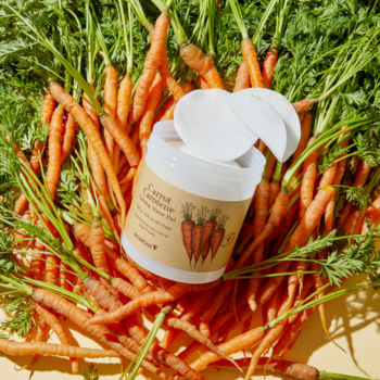 Biały słoik Carrot Carotene Daily Toning Pad z płatkami, otoczony świeżymi marchewkami, widok z góry.