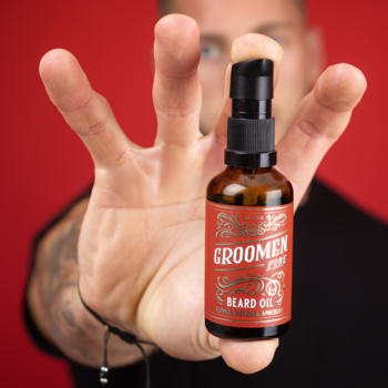 Męska dłoń trzyma brązową butelkę GROOMEN Beard Oil z czarną pompką na czerwonym tle, widok z bliska.