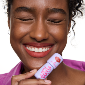 Uśmiechnięta kobieta z matowymi ustami trzyma fioletową pomadkę essence JUICY MATTE 01 Maple Rouge.