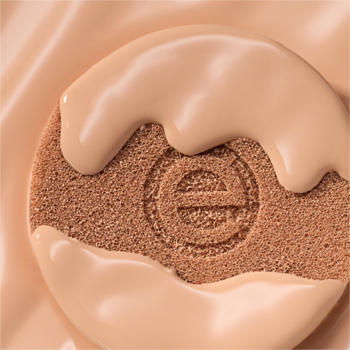 Beżowy podkład essence silky CUSHION 115, kremowy, spływający po brązowej poduszeczce z logo, zbliżenie.