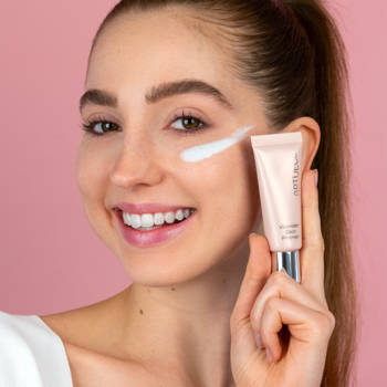 Uśmiechnięta kobieta nakłada bazę Artdeco Wonder Skin Primer na policzek. Beżowa tubka, efekt blur, różowe tło.