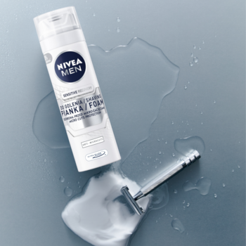 Biała pianka do golenia NIVEA MEN Sensitive z niebieskim logo, leży na mokrym szarym tle, obok maszynki i piany.