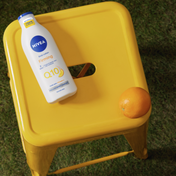 Biała butelka Nivea Q10 + witamina C ujędrniającego balsamu na żółtym taborecie obok pomarańczy, na trawie.