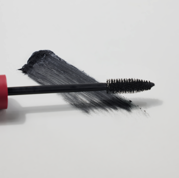 Zbliżenie na czarną szczoteczkę pogrubiającej maskary, która rozdziela rzęsy, rozmazany ciemny tusz na białym tle.
